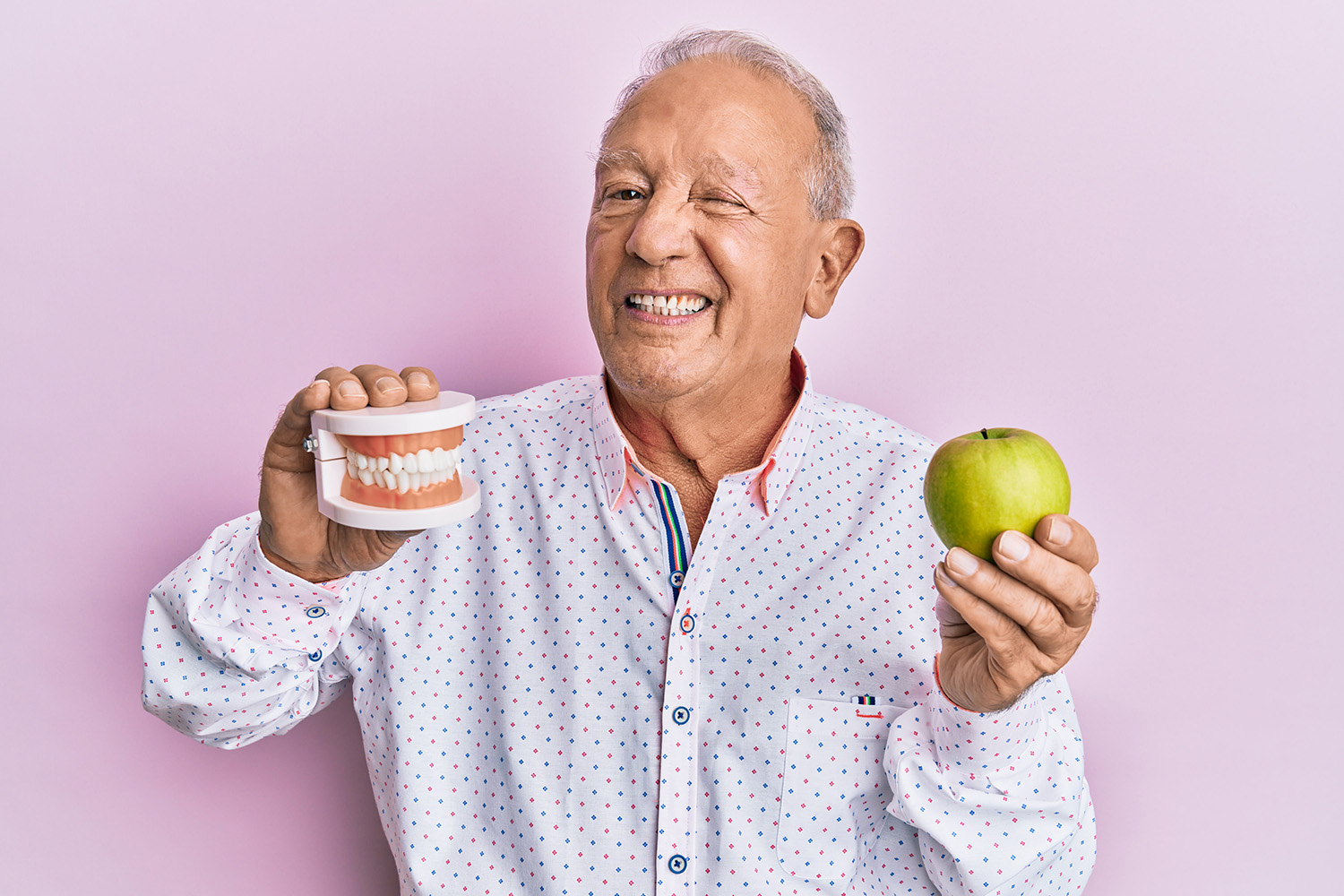 Senior caucasian man holding denture and green apple winking looking at the camera with sexy expression, cheerful and happy face.