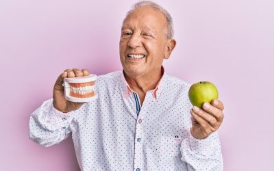 Senior caucasian man holding denture and green apple winking looking at the camera with sexy expression, cheerful and happy face.