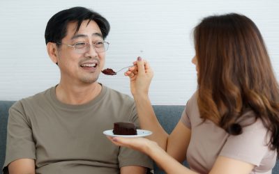 Portrait shot of cute smiling middle-aged Asian lover couple sitting on a couch at home, looking at each other with romantic feeling and eating a chocolate cake. Wife feeding her husband with care
